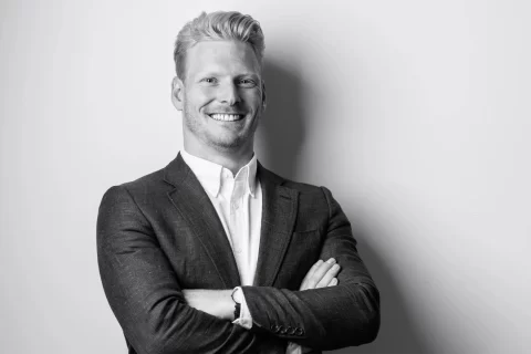 Black and white image of a smiling man with short, light-colored hair, wearing a dark suit jacket over a button-up shirt. Steve Buck stands against a plain backdrop with his arms casually crossed in front of his chest.