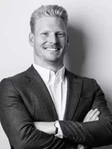 A black and white photo of Steve Buck, a smiling person with light-colored hair, wearing a blazer and a white dress shirt, with arms crossed in front of their chest. The background is plain, focusing on Steve Buck's facial expression and professional attire.