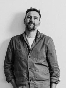 Black and white portrait of Steve Kennedy, a man with short hair and a beard, standing against a plain background. He is wearing a casual jacket with pockets and a t-shirt underneath. His hands are relaxed in his pockets as he looks slightly off camera with a calm expression.