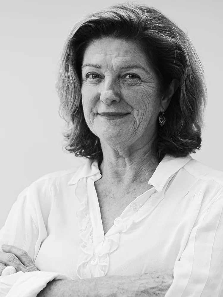 A black-and-white portrait of an older woman, Suellen Fitzgerald, with shoulder-length hair and a relaxed smile. She is wearing a light-colored blouse and has her arms gently crossed. The background is plain and light.