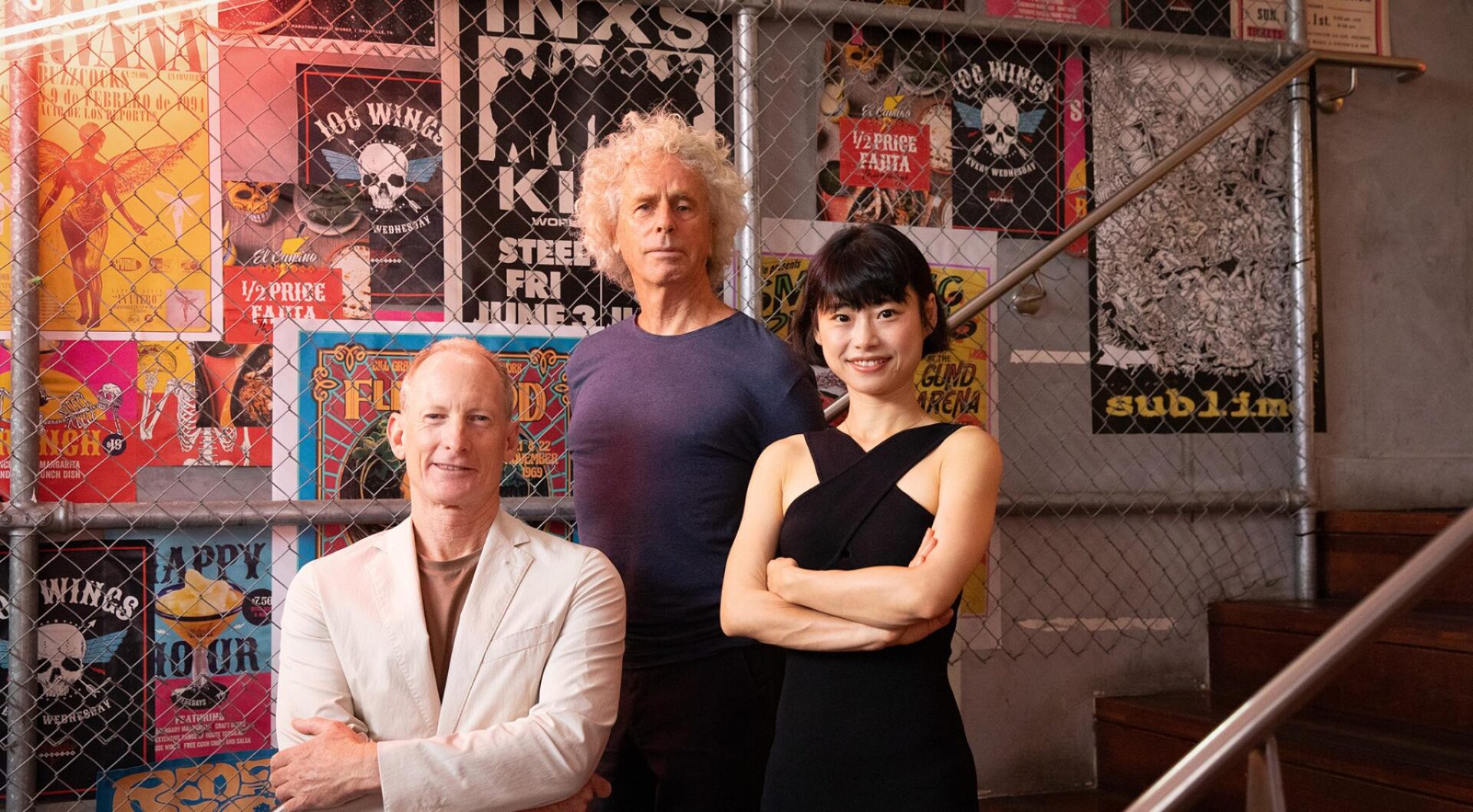 Three people are posing in front of a wall decorated with various colorful posters. On the left is McGregor Coxall founder and Chief Design Officer Adrian McGregor, he is standing on a lower stair, in the middle stands McGregor Coxall founder and Director of Design, Philip Coxall with curly blonde hair wearing a dark blue shirt, and on the right part-owner Ann Deng with black hair in a black dress.