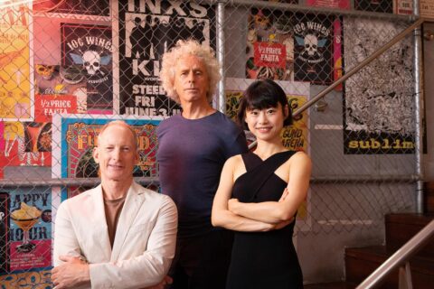 Three people are posing in front of a wall decorated with various colorful posters. On the left is a seated person in a white jacket, in the middle stands Director Ann Deng with curly blonde hair wearing a dark blue shirt, and on the right is a person with black hair in a black dress.