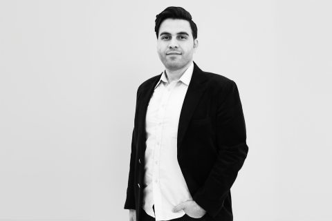 A black and white photo of Dr. Hossein Rizeei standing against a plain white background. Dressed in a dark blazer over a light-colored shirt, he has one hand in his pocket and is looking directly at the camera with a neutral expression.