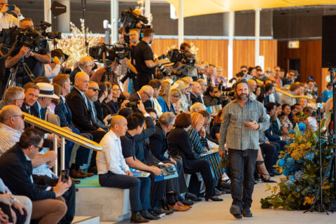 A crowd of people is seated in tiered rows in an outdoor setting, attentively observing a man walking in front of them. Several individuals are holding cameras and video equipment, indicating a media presence. The background features a modern, wooden structure, suggesting an awards ceremony ambiance.