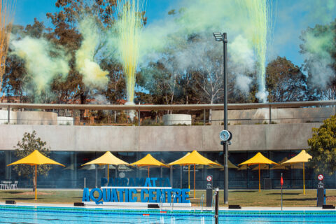 The Parramatta Aquatic Centre features wide, clear blue swimming pools in the foreground. In the background, yellow smoke and fireworks are set off above the modern, concrete buildings and shaded areas with bright yellow canopies, creating a festive atmosphere worthy of awards.