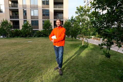 Logan Pennington, wearing a bright orange jacket and blue jeans, stands on a grassy area in front of an apartment building. Holding a white object and smiling, they are framed by trees and lush greenery in the background.