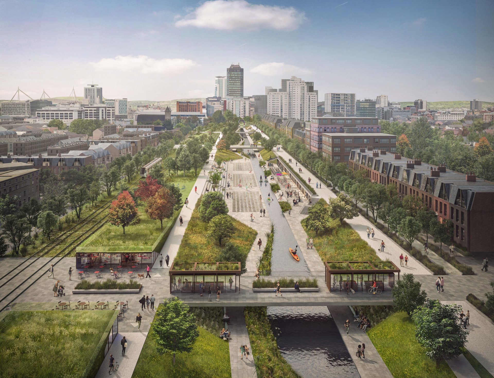 A large urban park with pathways, green spaces, and waterways runs through the middle of Cardiff City, flanked by buildings and trees. People are walking, biking, and sitting on benches. In the distance, high-rise buildings near the bay are visible against a blue sky with scattered clouds.