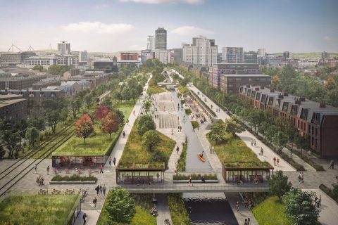 A large urban park with pathways, green spaces, and waterways runs through the middle of Cardiff City, flanked by buildings and trees. People are walking, biking, and sitting on benches. In the distance, high-rise buildings near the bay are visible against a blue sky with scattered clouds.