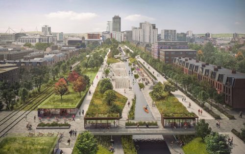 A large urban park with pathways, green spaces, and waterways runs through the middle of Cardiff City, flanked by buildings and trees. People are walking, biking, and sitting on benches. In the distance, high-rise buildings near the bay are visible against a blue sky with scattered clouds.