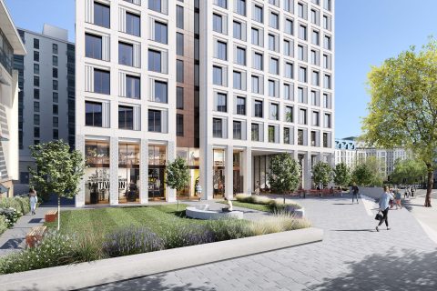 A modern multi-story building with large windows, surrounded by trees and landscaping in the bustling Haymarket area. There are people walking and sitting on benches in a paved area in front. The ground level features commercial spaces with storefronts. Clear blue sky in the background.