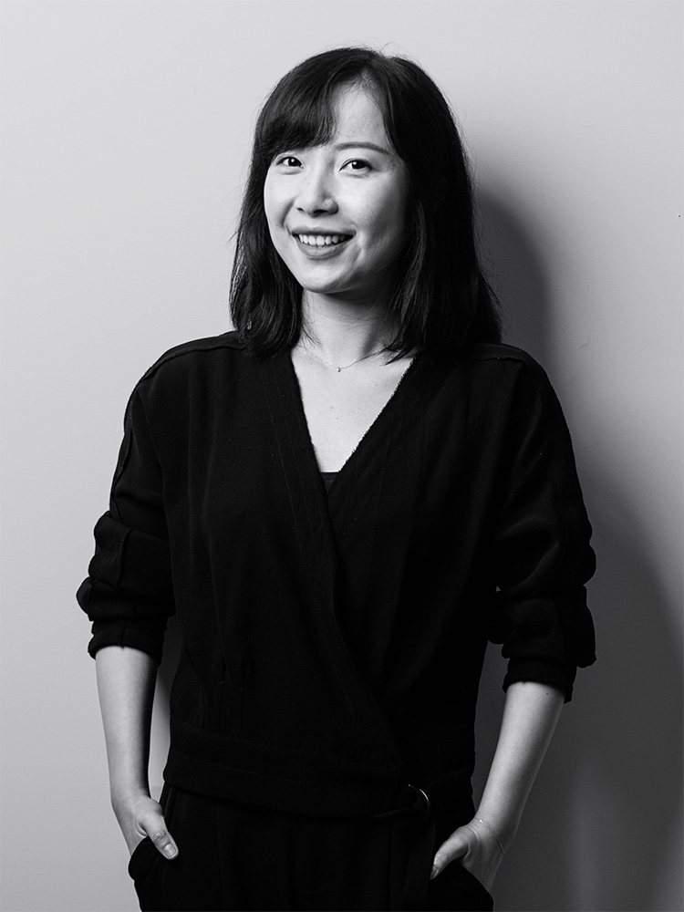 Black and white photo of Amber Guo, a young woman with shoulder-length hair, smiling and standing against a plain background. She is wearing a dark, long-sleeved top and has her hands in her pockets.