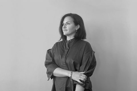 A black and white photo of Ana Abram, with shoulder-length dark hair, wearing a loose, long-sleeved shirt. She stands against a plain background, looking slightly away from the camera with a calm expression, one arm crossed over her body.