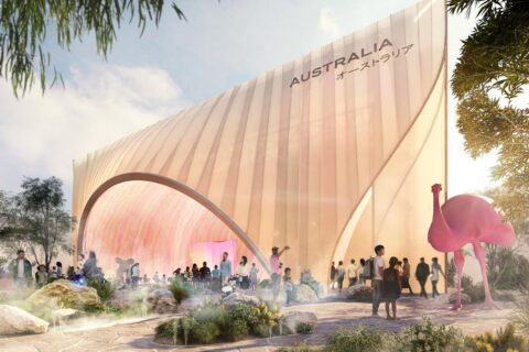 The Australian Pavilion at Expo 2025 in Osaka, Japan boasts a sleek, curved design with "Australia" written in English and Japanese on the facade. People gather outside the modern structure, where a large, stylized pink bird sculpture stands prominently amid lush greenery.