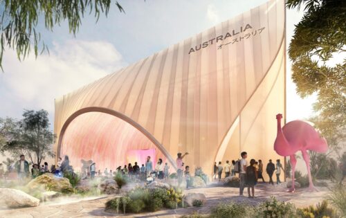 The Australian Pavilion at Expo 2025 in Osaka, Japan boasts a sleek, curved design with "Australia" written in English and Japanese on the facade. People gather outside the modern structure, where a large, stylized pink bird sculpture stands prominently amid lush greenery.