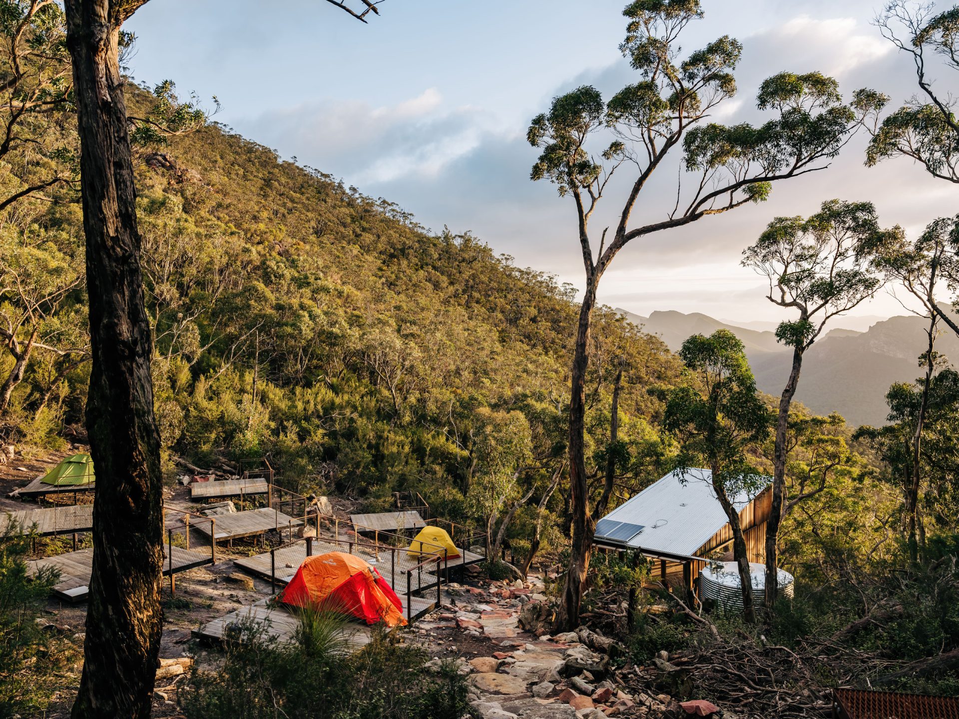 A group of tents is pitched on rocky, terraced platforms in a wooded, mountainous area at sunset. A small building with a metal roof is visible among the trees, and the Grampians Peaks Trail stretches out toward the horizon, offering breathtaking views of Gariwerd.