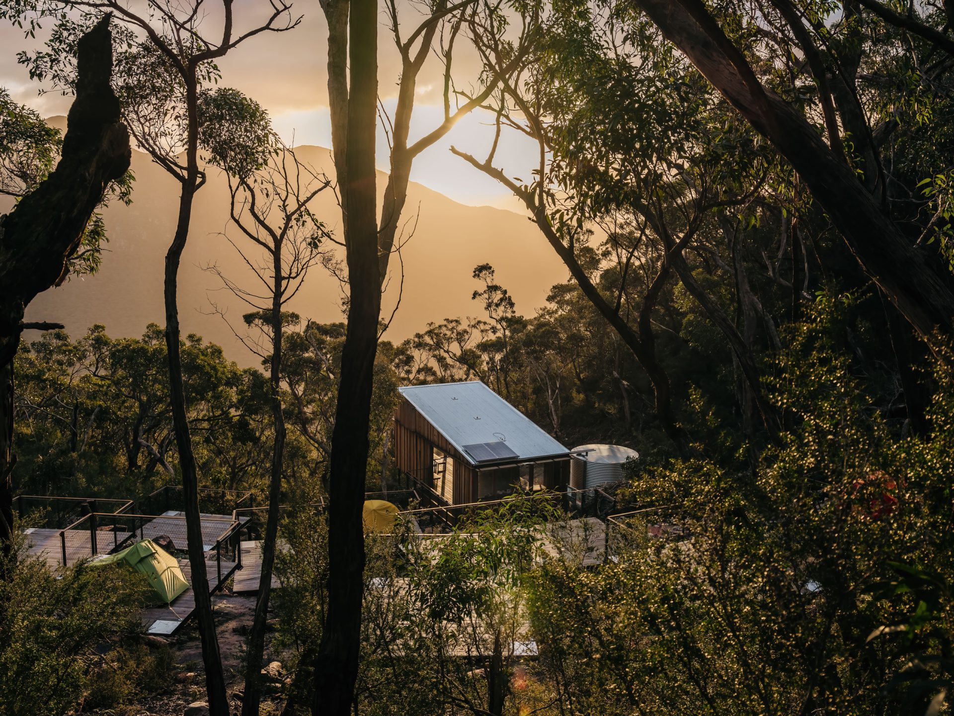 A small, secluded cabin with a metal roof is nestled among dense trees on a mountainside at sunset in Gariwerd. Soft golden light filters through the tree branches, illuminating the cabin and surrounding area. A deck extends from the cabin, and a green tent is pitched nearby along the Grampians Peaks Trail.