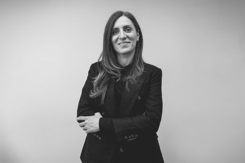 Black-and-white photo of Dr Behnaz Avazpour with long hair, wearing a dark blazer and shirt, standing against a plain background. They have a slight smile and their arms are crossed confidently.