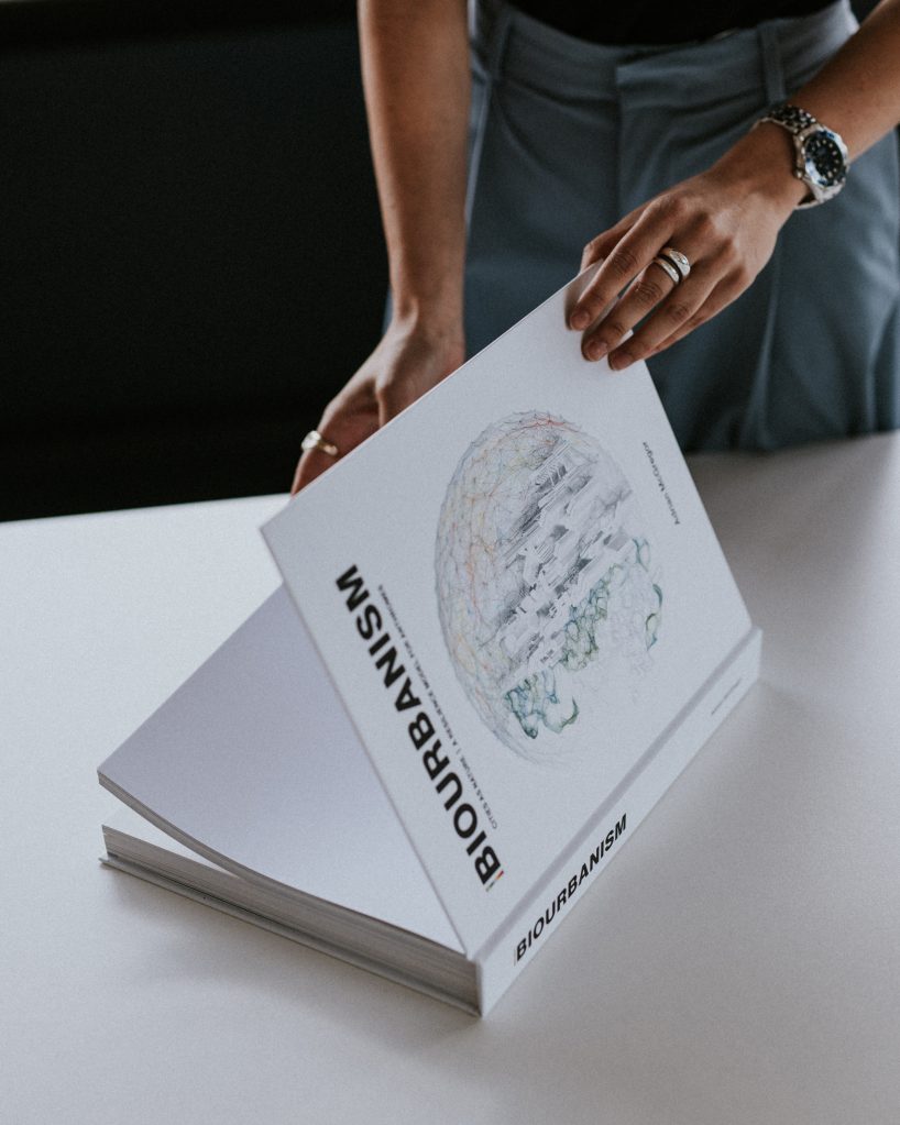 A person wearing a watch and rings is seen flipping through a large book titled "BIOURBANISM" at home. The open book displays an intricate illustration of a globe. The person is standing at a white table in a well-lit space.