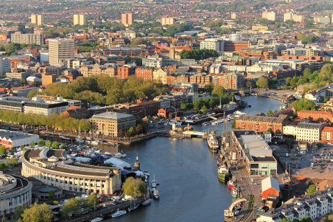 Aerial view of the bustling harbor city of Bristol, featuring a river that winds through the center, lined with boats, buildings, and lush green trees. Numerous buildings, both historical and modern, are visible with high-rise structures scattered throughout the background.