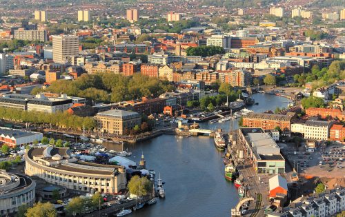 Aerial view of the bustling harbor city of Bristol, featuring a river that winds through the center, lined with boats, buildings, and lush green trees. Numerous buildings, both historical and modern, are visible with high-rise structures scattered throughout the background.