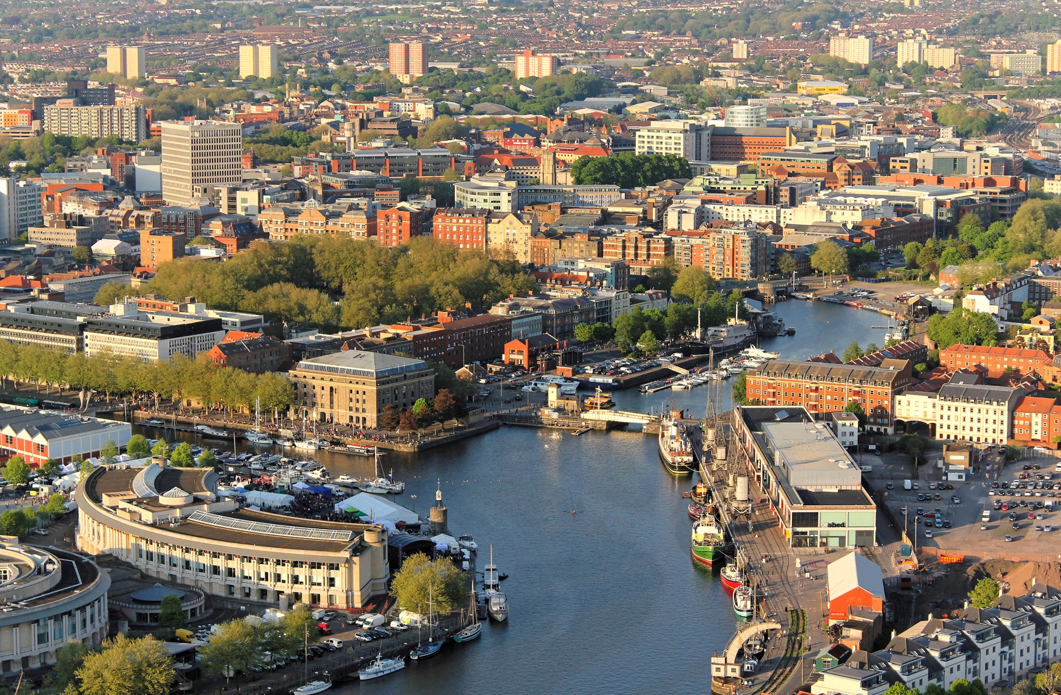 Aerial view of the bustling harbor city of Bristol, featuring a river that winds through the center, lined with boats, buildings, and lush green trees. Numerous buildings, both historical and modern, are visible with high-rise structures scattered throughout the background.