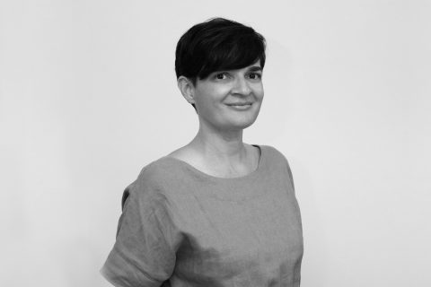 Black and white photo of Clare Mayberry with short dark hair, wearing a light-colored top. They are smiling softly and standing against a plain, light-colored background.