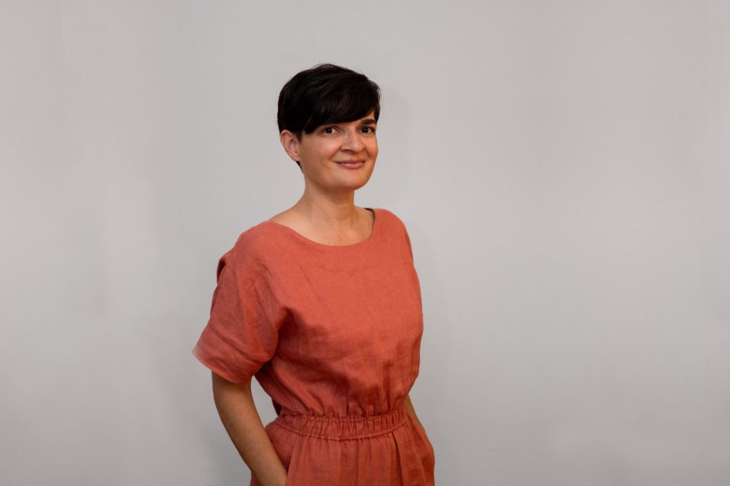 Clare Mayberry, with short dark hair, stands against a plain gray background wearing a rust-colored dress. Smiling and with one hand in her pocket, she exudes an effortless charm.