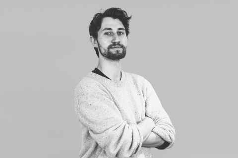 Black and white image of Daniel Ichallalene standing with his arms crossed. He has dark hair, a beard, and is wearing a light-colored, long-sleeve sweater. The background is a solid, light gray color.