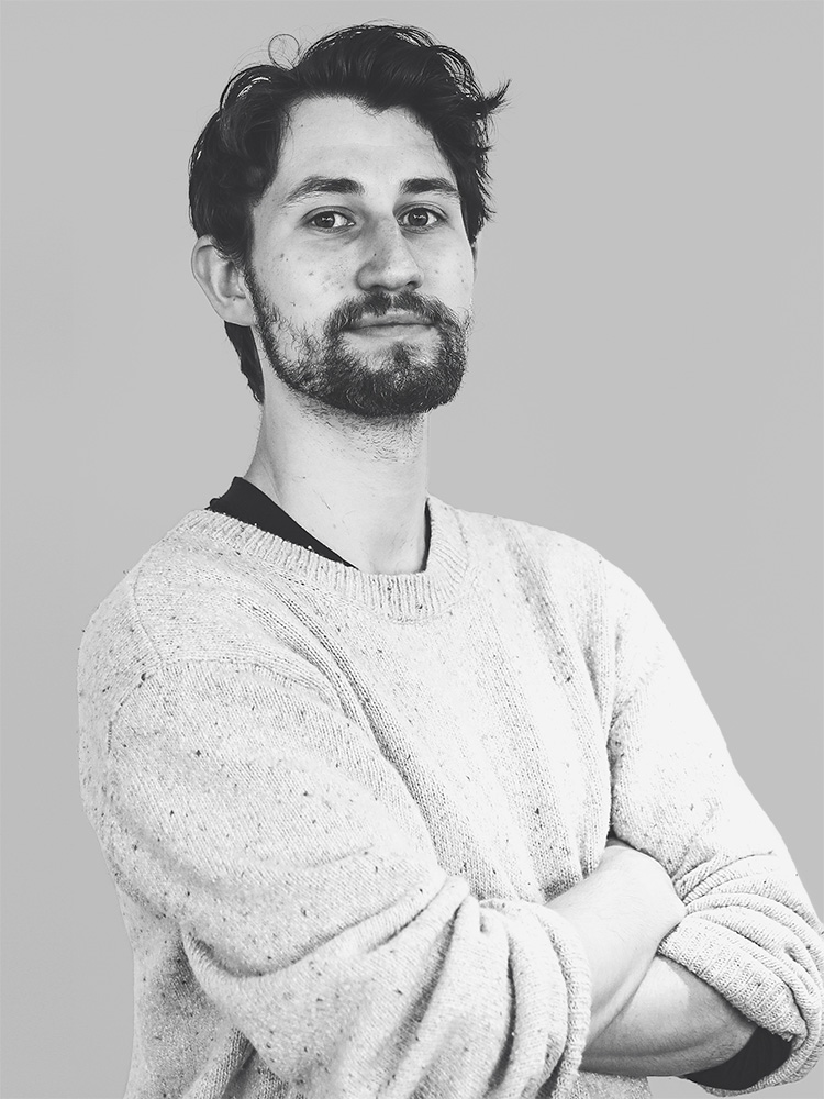 A black and white photo of Daniel Ichallalene, a bearded person with short, wavy hair, standing against a plain backdrop. They are wearing a light-colored crewneck sweater and have their arms crossed, with a slight smile on their face.