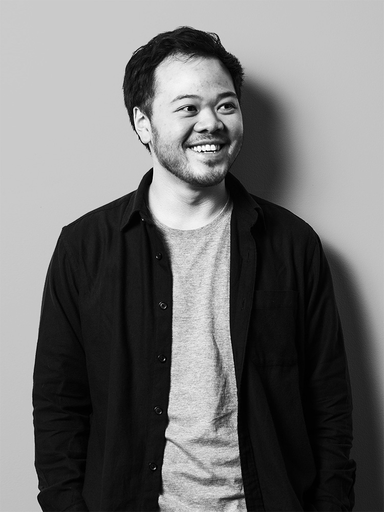 Black and white image of Daryl Wong with short dark hair and a trimmed beard, smiling and looking slightly to the side. They are wearing a light-colored t-shirt underneath a dark button-up shirt. The background is plain and light-colored.