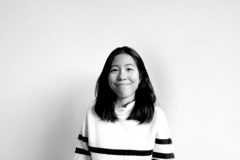 A person with long, dark hair and a slight smile stands against a plain white background. They are wearing a light-colored sweater with dark horizontal stripes on the sleeves. The image, capturing Rae Kwong's serene presence, is in black and white.