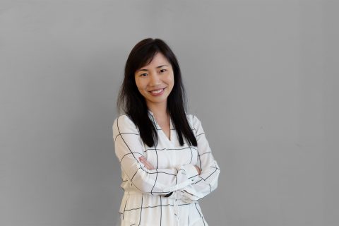 Dr. Elina Wu, with long dark hair, stands against a plain gray background dressed in a white blouse with a black grid pattern. She is smiling and has her arms crossed, exuding a confident and approachable demeanor.