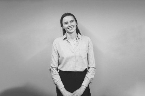 A black and white image of Freya Alston standing in front of a plain backdrop. She is wearing a light-colored button-up shirt and dark pants, with her hands loosely clasped in front. Freya is smiling and looking directly at the camera.