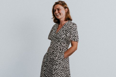 Gemma Hoult, a woman with shoulder-length hair, is wearing a black and white patterned dress as she stands against a plain white background. She is smiling and has her hands in the pockets of her dress.