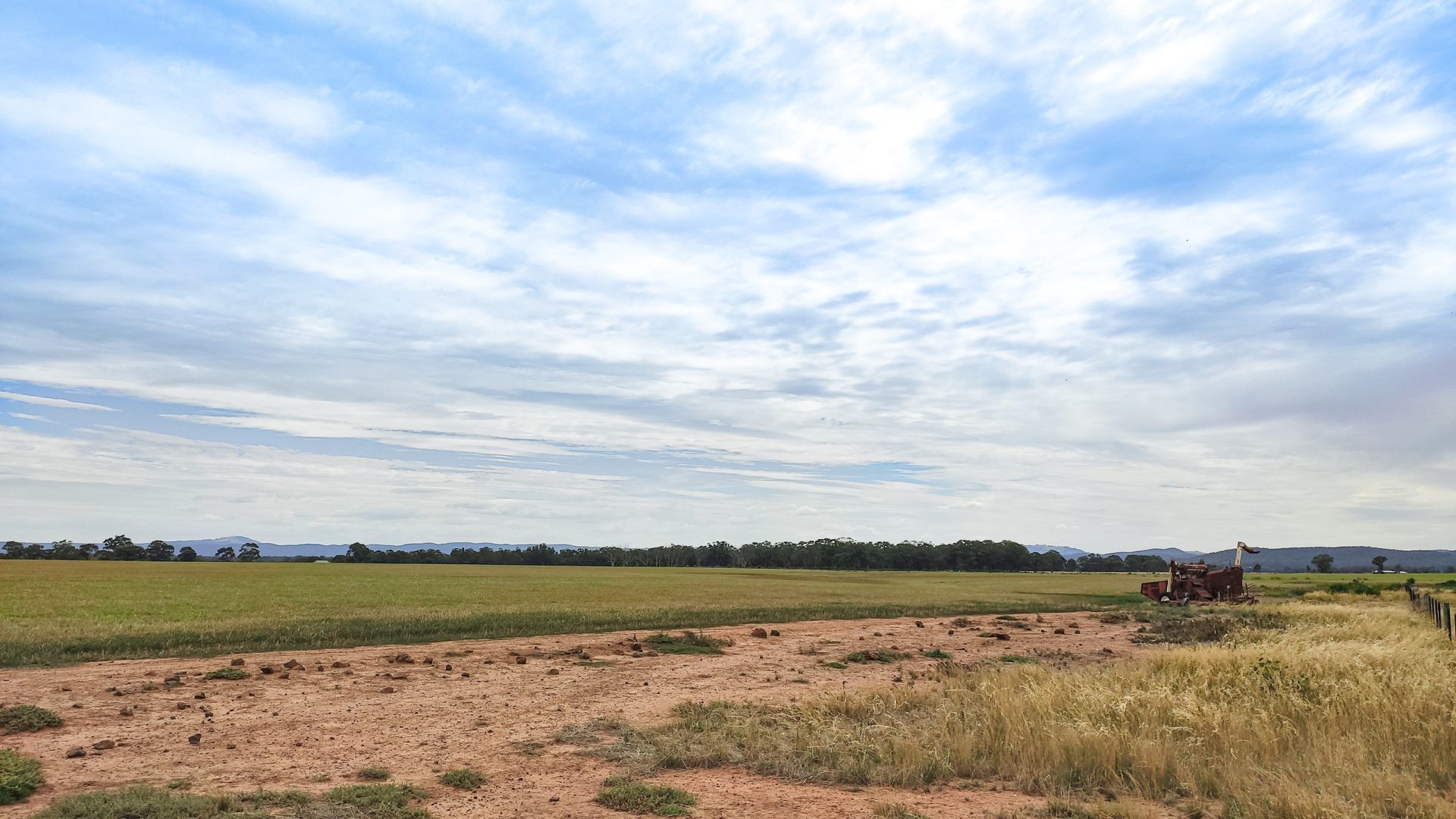 A vast open field stretches under a blue sky with scattered clouds. The ground in the foreground is a mix of bare patches and sparse grass. In the distance, trees line the horizon, and an old, rusted tractor sits to the right, close to the edge of what looks as if it could be part of a Harkness Cemetery masterplan.