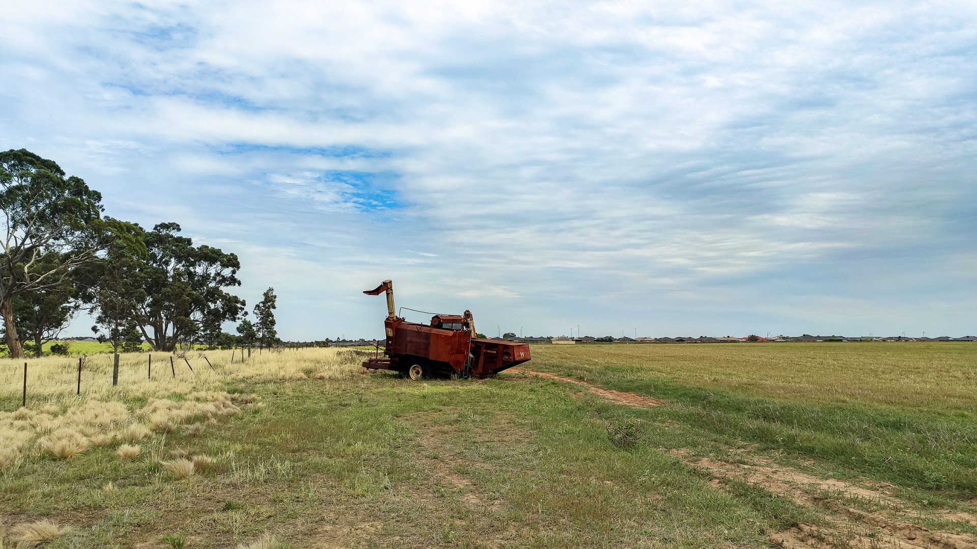 An old, rusted farm machine sits abandoned in a vast grassy field under a cloudy sky. A dirt path leads to the machine, with a wooden fence and tall trees on the left side of the image, reminiscent of Harkness Cemetery. Houses are visible in the distance on the horizon.