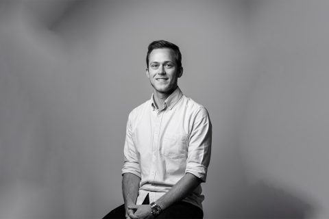 Black and white portrait of Jeremy sitting on a stool against a plain background. He is smiling, with short hair, wearing a button-up shirt with sleeves rolled up to his elbows, and has his hands resting on his thighs. The scene is simple and professional.