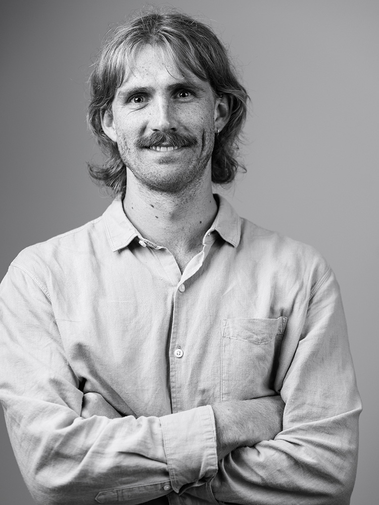 A black-and-white portrait of Logan Pennington with medium-length hair and a mustache, wearing a button-up shirt with rolled-up sleeves. They are standing against a plain background with their arms crossed and smiling slightly at the camera.