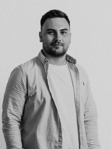 Black and white image of Luc Jones, a person with short, dark hair and a beard, standing and facing the camera. They are wearing a light-colored button-up shirt over a white t-shirt. The background is plain and light-colored.