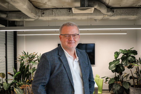 Leon Stone, McGregor Coxall CEO, stands in a modern office wearing glasses and a blazer, with plants and contemporary lighting in the background. A television screen and other office furniture are also visible.