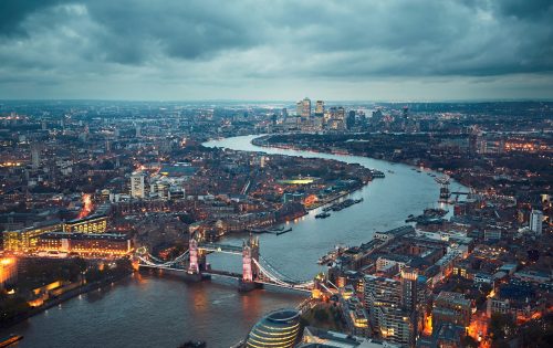 Aerial view of London at dusk, showcasing the illuminated Tower Bridge spanning the River Thames. The cityscape, featuring a mix of historic buildings and modern skyscrapers, highlights Transport for London's bustling network under a cloudy sky. The river winds through the city, reflecting the urban lights.