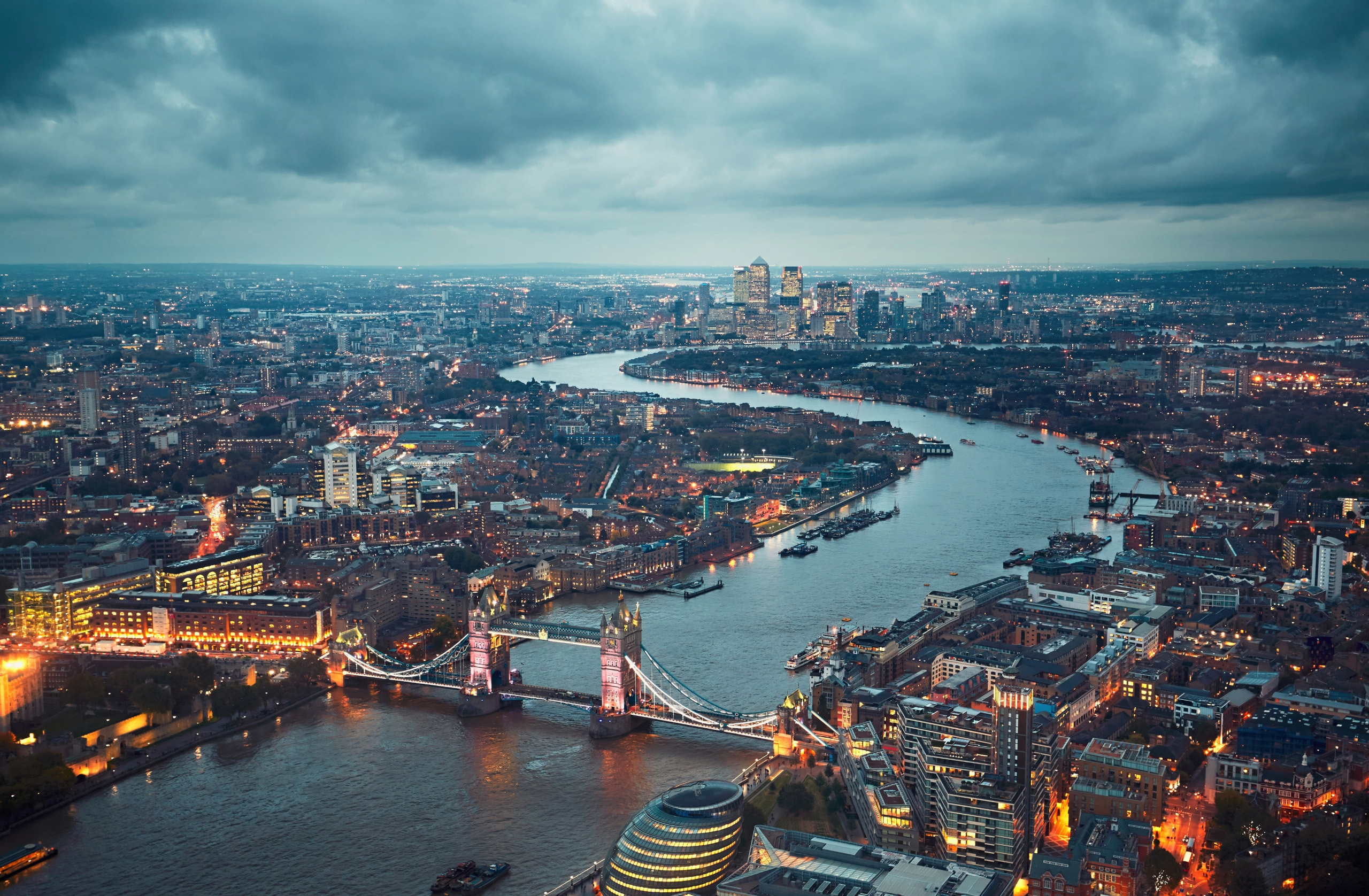 Aerial view of London at dusk, showcasing the illuminated Tower Bridge spanning the River Thames. The cityscape, featuring a mix of historic buildings and modern skyscrapers, highlights Transport for London's bustling network under a cloudy sky. The river winds through the city, reflecting the urban lights.