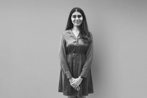 A woman with long, dark hair, wearing a knee-length, long-sleeved dress, stands against a plain background. She is smiling and has her hands clasped in front of her. The image of Manasi Sabnis is in black and white.