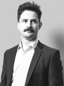 A black and white photograph of a man named Matthew Moore with short hair and a mustache. He is wearing a light-colored dress shirt and a dark suit jacket. Matthew gazes slightly to the left with a neutral expression on a plain background.