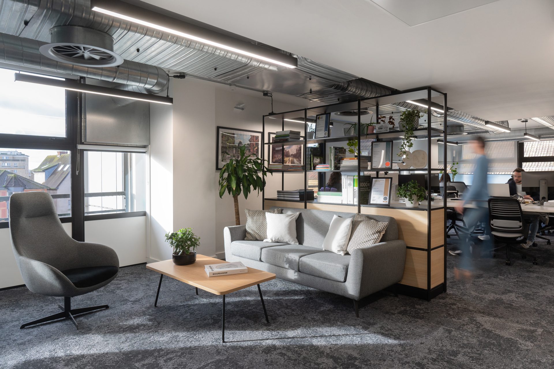 A modern office space featuring a seating area with a gray couch, a wooden coffee table adorned with plants, a gray armchair, and a shelf with books and decor. A person walks by in the background. The office, perfect for careers-focused activities, boasts large windows, exposed ducts, and ceiling lights.