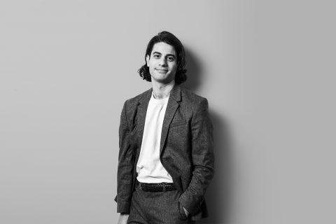 Black-and-white image of Michael Polifrone with shoulder-length hair standing against a plain background. He is wearing a suit with a white t-shirt underneath, smiling slightly with one hand in his pocket.