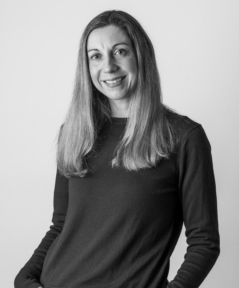 Black and white portrait of Jennifer McLoughlin with straight, long hair, wearing a dark, long-sleeved shirt. She is smiling and looking at the camera against a plain background, with one hand casually placed in her pocket.