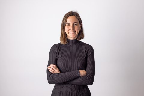 A person with shoulder-length hair, Asli Arda Healy-Smith, is smiling at the camera. They are wearing a long-sleeved black turtleneck and have their arms crossed. The background is plain white.