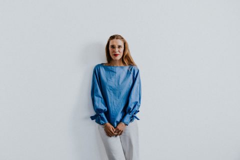 Sophie Thomas, with long blonde hair, stands against a plain white background. She is wearing a blue blouse with puffy sleeves tied at the wrists and white pants. Sophie looks directly at the camera with a slight smile.