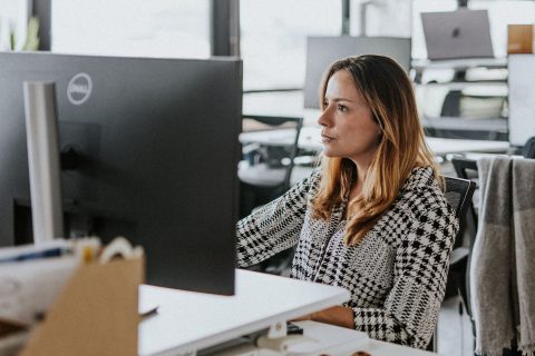 Camila Vidal, a woman with long hair, is sitting at a desk, focused on the large computer monitor in front of her. She is wearing a black and white patterned top in an office setting with other desks and computers in the background. A pair of sunglasses sits on the desk.